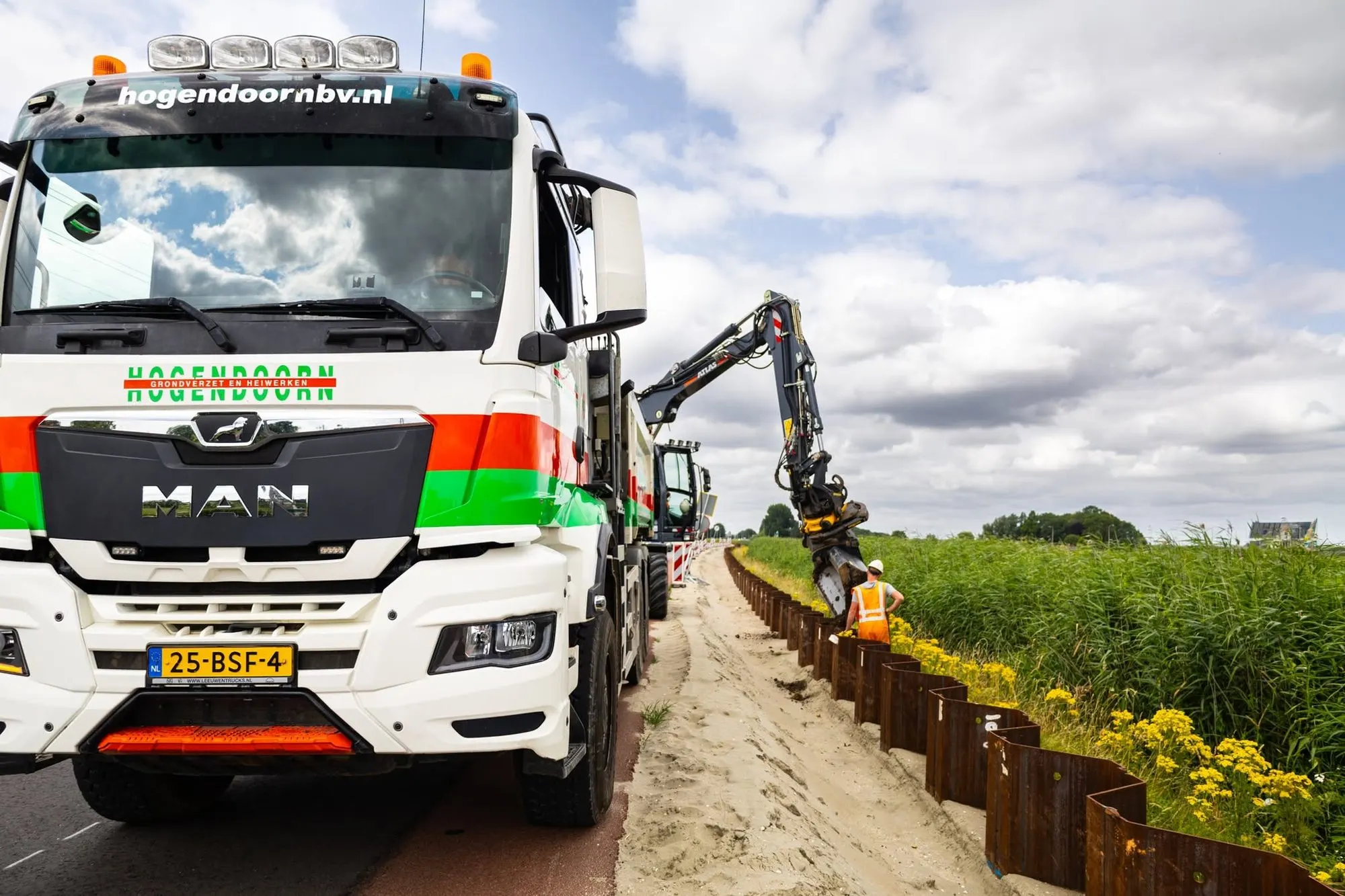 MAN kipper met Hogendoorn-belettering langs een dijk, mobiele kraan plaatst damwand