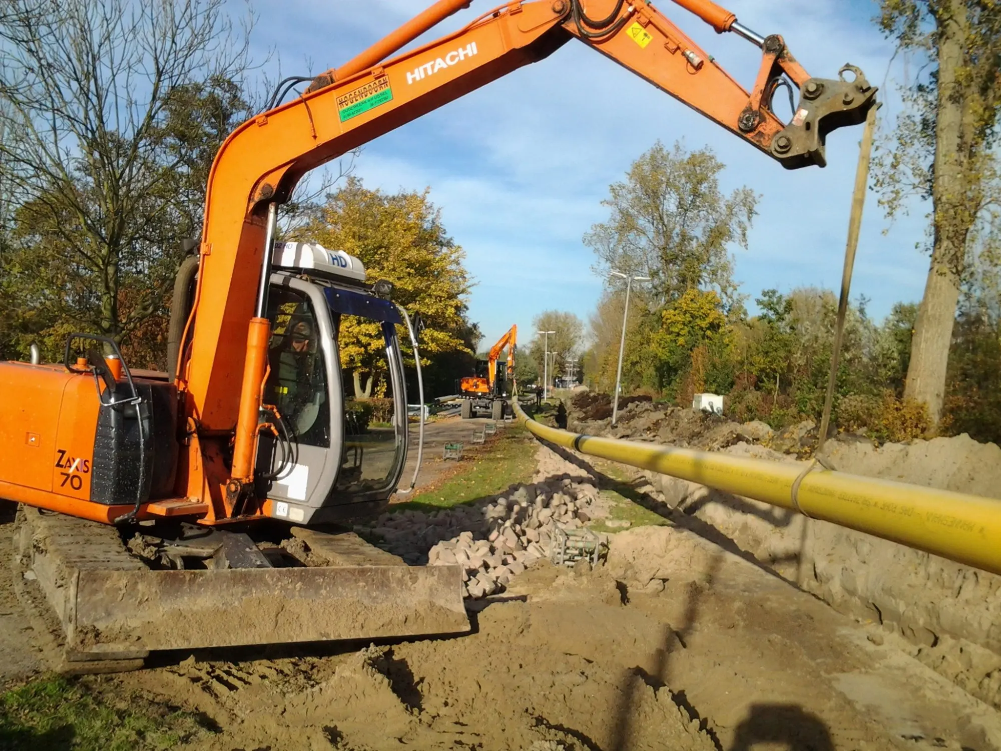 Hitachi ZX70 graafmachine graaft een sleuf voor een gele gasleiding langs een weg