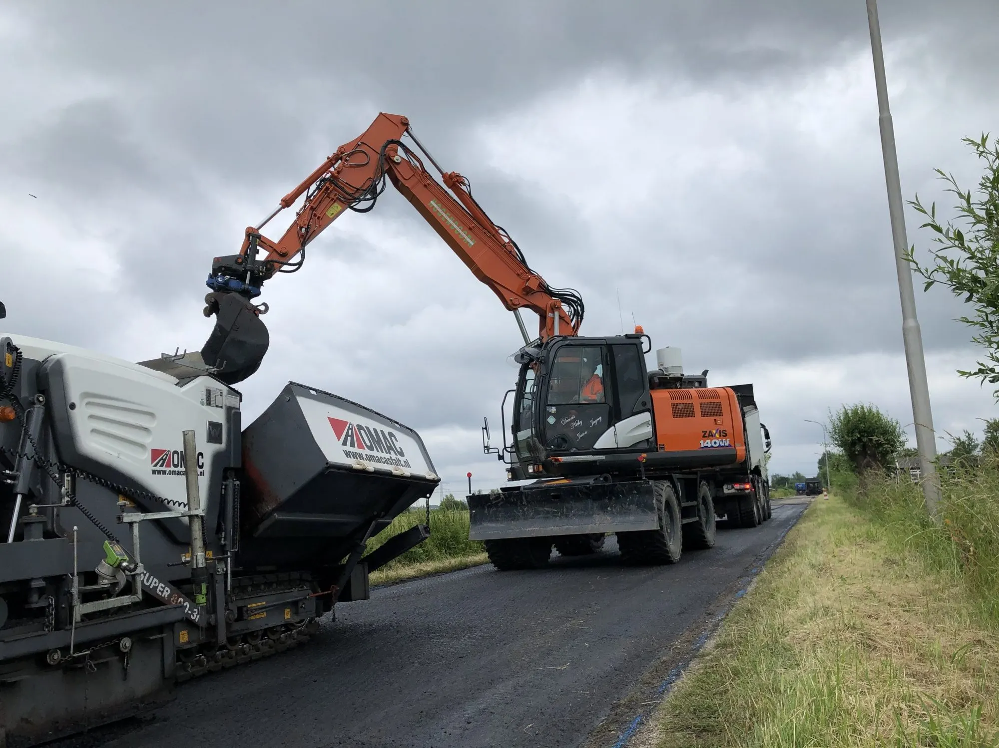 Hitachi ZX140W mobiele graafmachine vult een asfaltmachine op een dijkweg