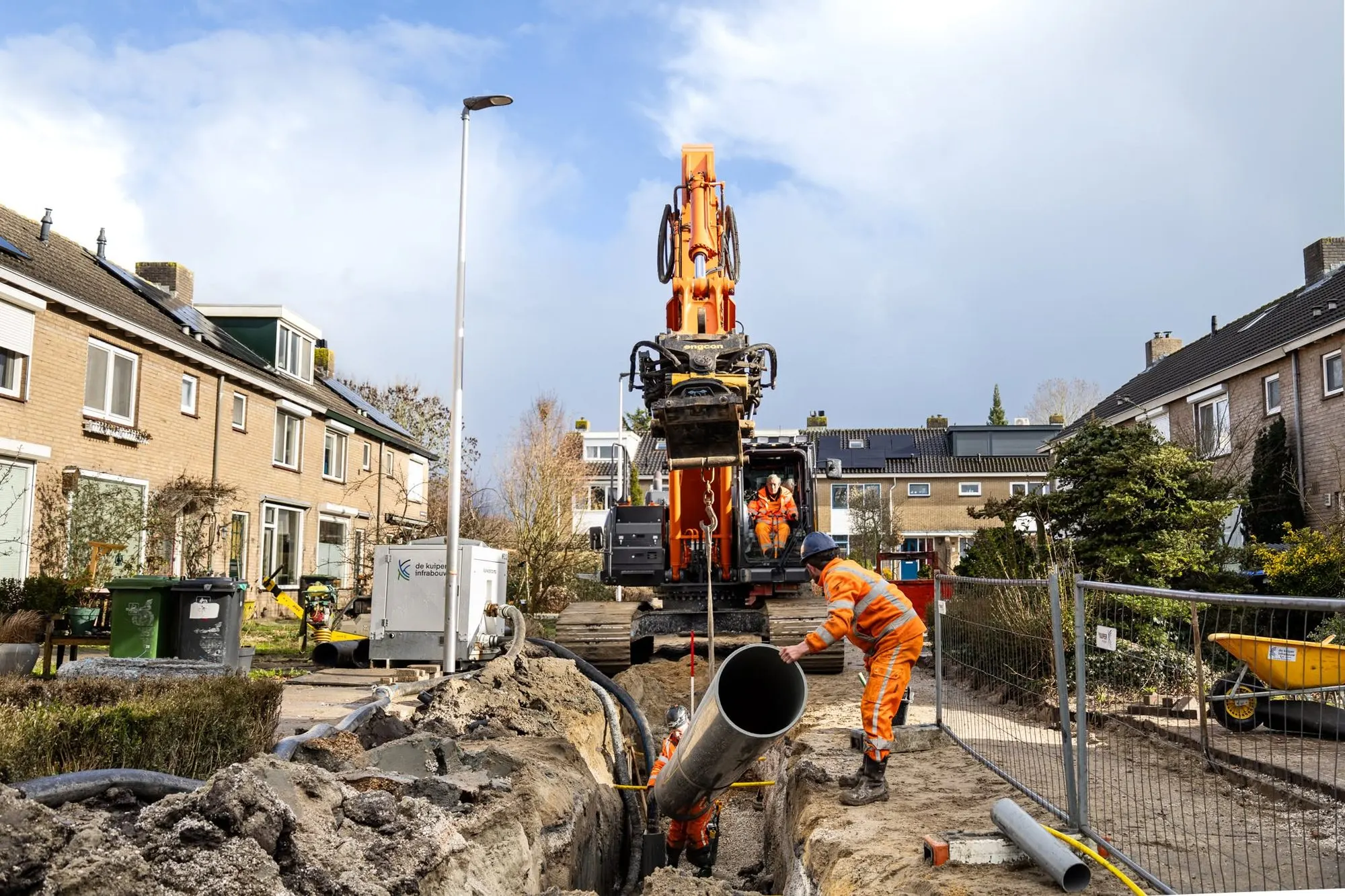 Oranje Hitachi rupskraan laat een rioolbuis in een sleuf zakken in een woonwijk