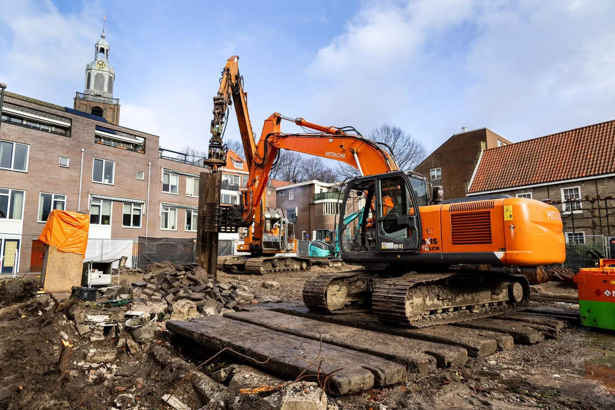 Twee oranje Hitachi rupskranen met trilblok bij damwandplanken, kerktoren op de achtergrond