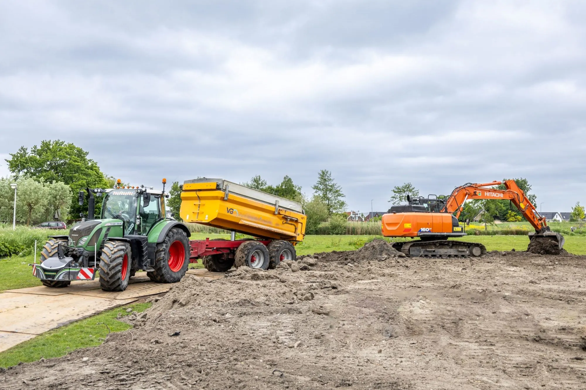 Fendt tractor met gele kieper en Hitachi rupskraan bij grondwerk op een grasveld