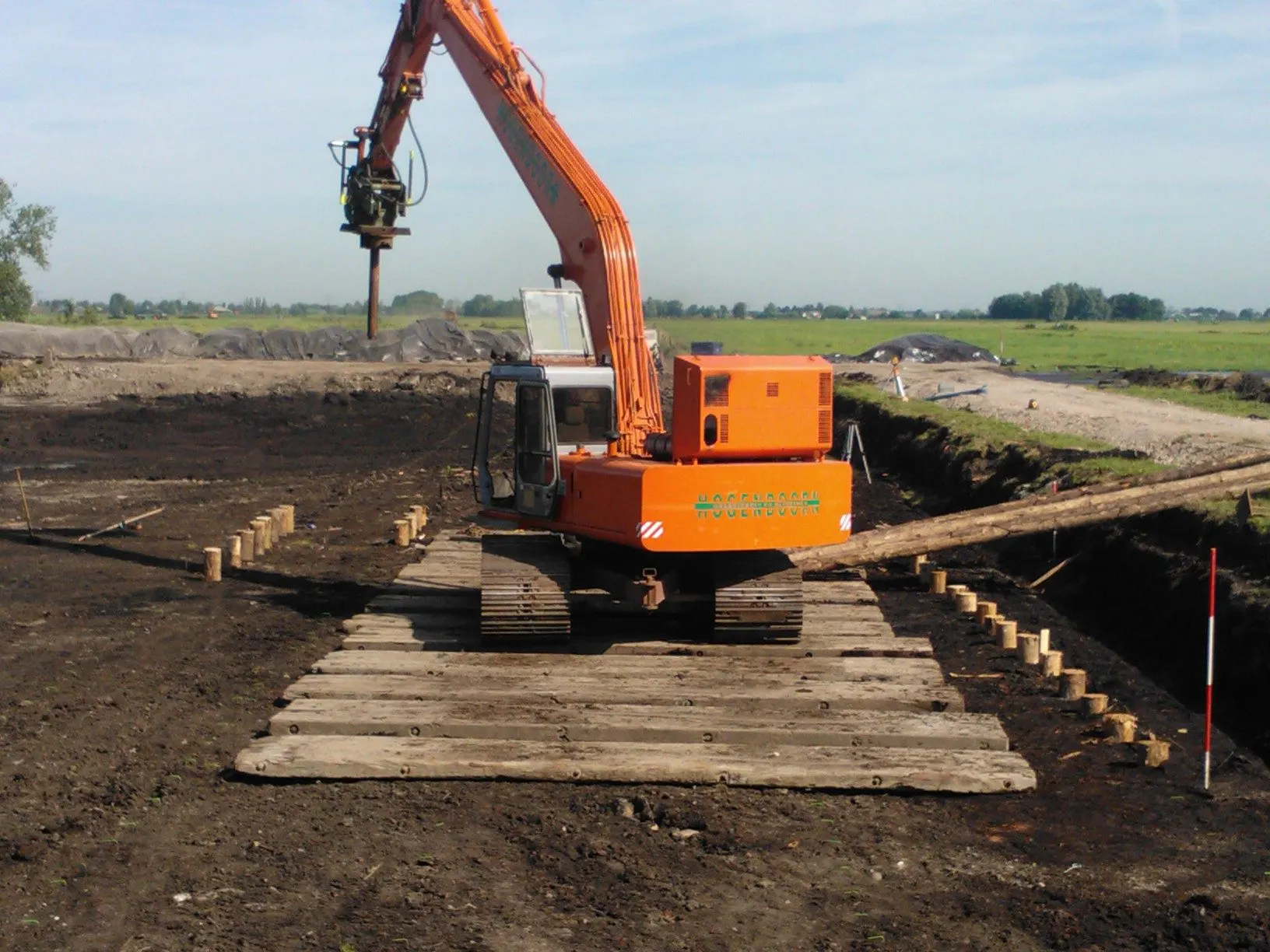 Rupskraan met trilblok op rijplaten bij houten palen
